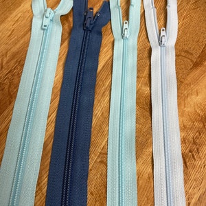 May include: Four zipper closures in different colors. Two are light blue, one is a darker blue, and one is a lighter shade of blue. The zippers are all closed.
