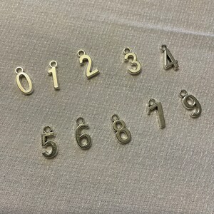 May include: Ten silver metal number charms, each with a different number from 0 to 9. The charms are arranged in two rows on a light gray textured surface.