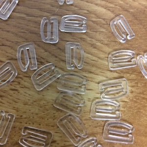 May include: A close-up of a pile of clear plastic bra strap adjusters. The adjusters are shaped like a letter 'D' with a small loop on the top.