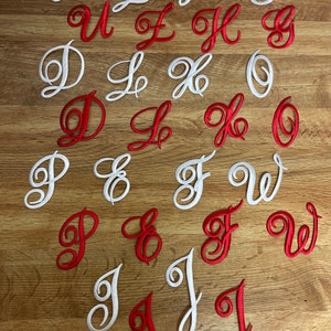 May include: A set of white and red embroidered letters in a cursive font. The letters are arranged in two rows on a wooden surface. The letters are U, L, H, G, D, X, 0, P, E, F, W, J.