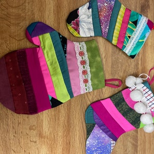 May include: Three colorful patchwork Christmas stockings with different embellishments. One stocking has a white lace trim and red buttons, another has a white and blue checkered trim with white pom-poms, and the third has a green fabric with a white floral pattern.