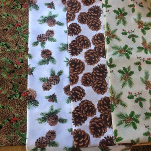 May include: Three fabric swatches with different patterns of pine cones and holly. The first swatch is black with brown pine cones and red holly berries. The second swatch is white with brown pine cones and green holly leaves. The third swatch is beige with brown pine cones and green holly leaves.