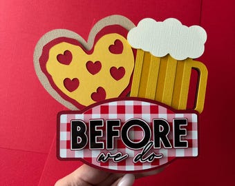 May include: A layered paper cutout decoration featuring a yellow heart with heart-shaped cutouts, a yellow beer mug with white foam, and a red and white checkered sign with the black text BEFORE we do, all arranged in a stacked design.