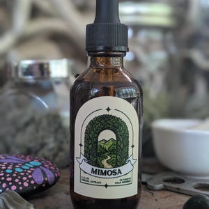 Puede incluir: Una botella de vidrio marrón con un cuentagotas negro. La etiqueta dice "Mimosa" con un diseño de arco verde. La botella contiene 60 ml de extracto de hierbas y es prensada en frío.