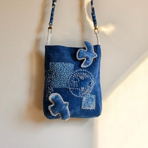 Peut inclure: Un sac bandoulière bleu avec une bandoulière tressée et des motifs cousus blancs. Le sac est orné de deux appliqués en forme d'oiseaux et de formes géométriques, le tout rehaussé de fil à broder blanc sur un fond en tissu bleu foncé.