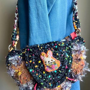 May include: Handmade black crochet bag with a colorful bunny applique. The bag features a textured design with colorful sequins and fringe. The strap is made of braided fabric and wooden beads.
