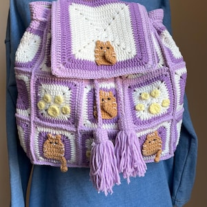 Peut inclure: Sac à dos crocheté fait main, design patchwork. Le sac présente des carrés blancs et lavande, avec des appliqués de chats et d'empreintes de pattes. Deux pompons lavande pendent à l'avant. Un accessoire unique et artisanal.