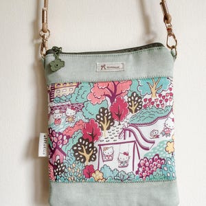 May include: A light green crossbody bag featuring a colourful print of Hello Kitty characters in a park with trees, a slide, and a swing set. The bag has a zip closure with a flower-shaped pull, a tan strap, and a tag that reads elflotus.