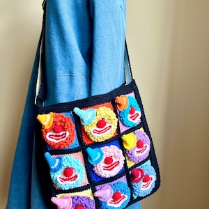 May include: A black crocheted shoulder bag with a patchwork design of clown faces. Each square features a different coloured clown face with a red nose, white mouth, and a small hat. The bag has a black strap and a blue background.