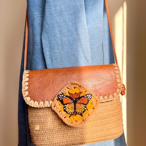 May include: A brown and tan crossbody bag with a woven straw body and a brown leather flap. A crocheted square with a butterfly design is centered on the front. The bag has a long, thin strap.