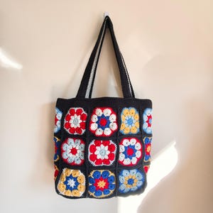 May include: A handmade crochet tote bag constructed from a series of square motifs featuring floral patterns in red, blue, yellow, and white. The squares are joined by a solid black yarn border, and the bag includes two matching black shoulder straps.