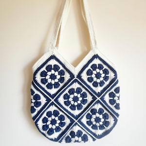 May include: A crochet tote bag constructed from multiple square motifs featuring a dark navy blue floral pattern on a white background. The bag includes two integrated white crochet shoulder straps and a V-shaped opening at the top centre.