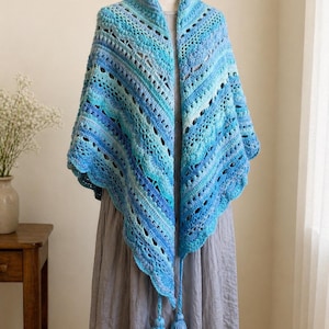 May include: A handmade, blue crochet shawl with a triangular shape. The shawl features a gradient of blue hues, from light to dark, with intricate openwork patterns. Two blue tassels hang from the bottom corners. The shawl is displayed on a mannequin.