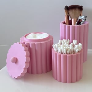 Puede incluir: Tres recipientes cilíndricos de color rosa con textura acanalada vertical y bordes festoneados. Un recipiente contiene bastoncillos de algodón blancos, otro discos de algodón y el tercero brochas de maquillaje y un rizador de pestañas.