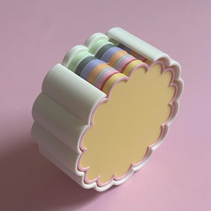 Op de afbeelding: Een witte container met geschulpte randen en pastelkleurige linten aan de binnenkant. De container heeft een gele binnenkant en een roze omtrek. De linten zijn in tinten groen, grijs, blauw, oranje en roze. De container staat op een roze achtergrond.