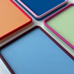 Puede incluir: Cuatro bandejas rectangulares con bordes elevados, cada una con una combinación de colores distinta: una bandeja azul con borde rosa, una naranja con borde rojo, una verde claro con borde magenta y una azul claro con borde morado oscuro.