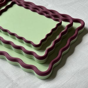 May include: A set of three rectangular trays stacked on top of each other. Each tray features a flat, light green surface with a raised, wavy, dark purple border. The trays are arranged in descending order of size, creating a layered effect.
