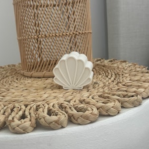 Könnte beinhalten: Ein weißes, muschelförmiges Dekorationsobjekt liegt auf einem gewebten, kreisförmigen Platzset. Im Hintergrund befindet sich eine hohe, gewebte Korblampe. Das Platzset hat einen dekorativen, geflochtenen Rand. Die Objekte befinden sich auf einer weißen Oberfläche.