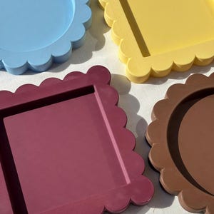 Puede incluir: Cuatro bandejas decorativas con bordes festoneados. Una bandeja circular azul claro, una bandeja cuadrada amarilla, una bandeja rectangular color granate oscuro y una bandeja circular marrón. Cada bandeja tiene un acabado mate y un centro hundido.