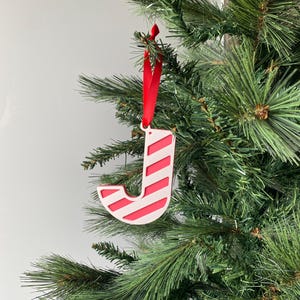 May include: A festive Christmas tree decoration in the shape of the letter 'J'. The white ornament features red diagonal stripes and is suspended by a red ribbon. It hangs on a green Christmas tree.