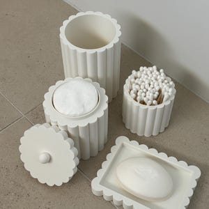 Puede incluir: Un conjunto de accesorios de baño blancos con un diseño de bordes festoneados. La colección incluye un recipiente cilíndrico alto, un contenedor pequeño con tapa lleno de discos de algodón, un frasco abierto con bastoncillos y una jabonera rectangular.