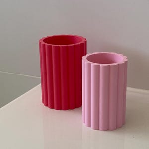 Op de afbeelding: Twee cilindrische decoratieve vazen. De ene is felrood, de andere zachtroze. Beide vazen hebben een gegroefd ontwerp en een geschulpte bovenrand. Ze staan op een wit oppervlak.