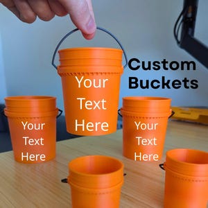 May include: Orange custom buckets of various sizes, with the text "Your Text Here" on some. The image also includes the text "Custom Buckets" and "Various sizes available!"