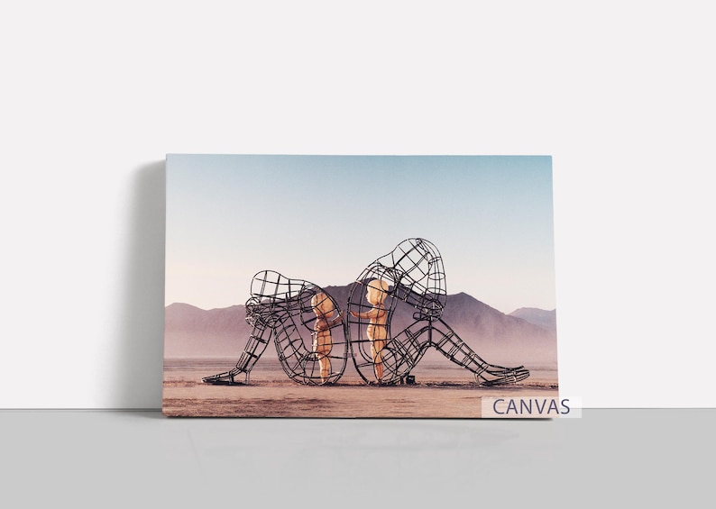 Pu&ograve; includere: Stampa su tela con due sculture a filo di figure sedute, schiena contro schiena, in un paesaggio desertico. Le sculture sono scure, con un interno chiaro. Montagne e cielo blu fanno da sfondo. La parola "CANVAS" &egrave; stampata in basso a destra.