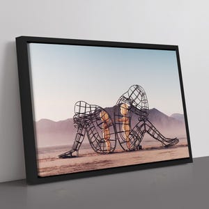 Pu&ograve; includere: Stampa artistica incorniciata raffigurante una scultura di due figure intrecciate, costruite con un design a filo metallico, ambientata in un paesaggio desertico con montagne e un cielo azzurro pallido. L'opera &egrave; incorniciata in nero.
