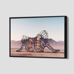 May include: Framed artwork featuring two wireframe human sculptures in a desert landscape. The sculptures are seated back-to-back, with one head resting on its knees. The background shows mountains and a pale blue sky.