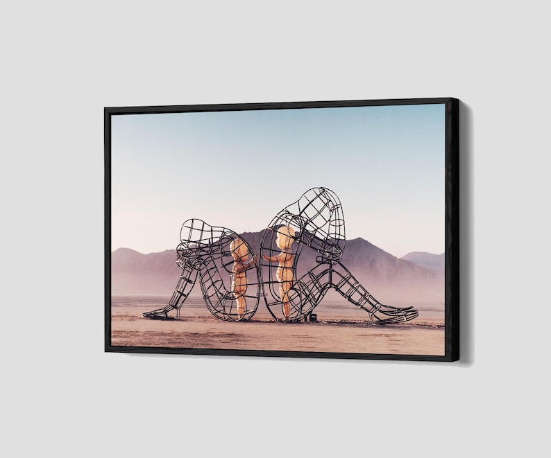 Pu&ograve; includere: Opera d'arte incorniciata con due sculture umane a filo metallico in un paesaggio desertico. Le sculture sono sedute schiena contro schiena, con una testa appoggiata sulle ginocchia. Lo sfondo mostra montagne e un cielo azzurro pallido.
