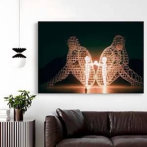 May include: A large canvas print depicts two wireframe figures facing each other, with a smaller figure between them. The figures are illuminated with a warm glow against a dark background. A person stands in front of the figures.