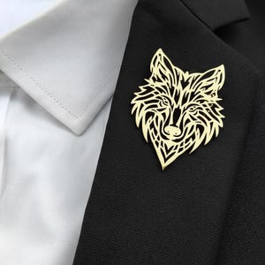 Può includere: Un fermaglio a forma di testa di lupo color oro appuntato su un blazer nero. Il fermaglio è una rappresentazione dettagliata e stilizzata del volto di un lupo, con intricati motivi ritagliati. Il blazer è abbinato a una camicia bianca con colletto.