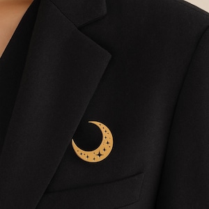 Puede incluir: Broche dorada con forma de media luna con detalles de estrellas, prendida a una blazer negra. El broche presenta un diseño recortado de estrellas, un accesorio de temática celestial.