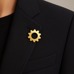 Peut inclure: Une épingle de revers dorée en forme de roue dentée est fixée à un blazer noir. L'épingle a un centre circulaire et des dents espacées régulièrement sur le pourtour. Le tissu du blazer est sombre et lisse.