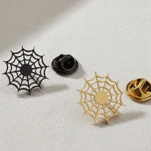 Könnte beinhalten: Zwei spinnennetzförmige Emaille-Anstecknadeln, eine schwarze und eine goldfarbene, mit passenden Nadelrücken. Die Anstecknadeln befinden sich auf einer strukturierten, cremefarbenen Oberfläche. Die goldene Anstecknadel hat ein glänzendes Finish, während die schwarze Anstecknadel matt ist.