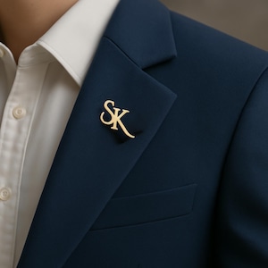 Może przedstawiać: Złota przypinka do klapy z inicjałami "SK" w ozdobnej czcionce jest przymocowana do granatowej marynarki. Marynarka jest noszona na białej koszuli. Przypinka to stylowy dodatek.