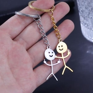 May include: Two stick figure keyrings, one silver and one gold, each with a smiley face and a raised middle finger. The keyrings are attached to a gold and silver keyring.