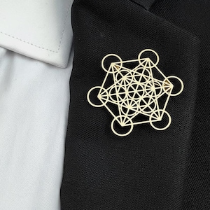 Peut inclure: Une épingle géométrique dorée, composée de cercles et de lignes interconnectés, est fixée sur un blazer noir. Le design de l'épingle est complexe et symétrique, créant un accessoire visuellement attrayant. Le blazer est associé à une chemise blanche à col.