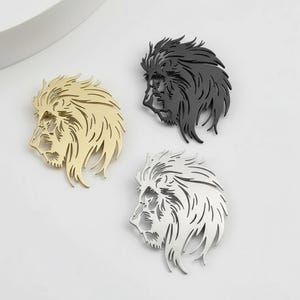 May include: Three metal lion head ornaments in gold, black, and silver. Each lion head is a profile view with detailed mane and facial features. The ornaments are likely for decorative purposes.