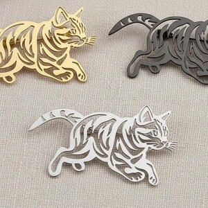 May include: Three metal cat brooches in gold, silver, and dark gray. Each brooch depicts a running cat with detailed stripes and whiskers. The brooches are designed with cut-out patterns, giving them a delicate appearance.