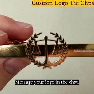May include: A gold tie clip with a scales of justice emblem within a laurel wreath. The text "Custom Logo Tie Clips" is at the top, and "Message your logo in the chat" is at the bottom. The clip is held by a person.