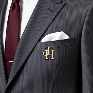 Puede incluir: Un traje de raya diplomática gris oscuro con una corbata burdeos y un pañuelo blanco. Un pin con monograma dorado está fijado a la solapa del traje. El traje está hecho a medida y parece estar hecho de una tela de alta calidad.
