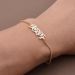 Puede incluir: Una pulsera dorada con una cadena fina y un dije rectangular con caracteres japoneses. El dije es el punto focal, con los caracteres prominentemente mostrados. La pulsera se lleva en una muñeca.