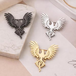 May include: Three phoenix brooches in black, silver, and gold. Each brooch features a detailed bird design with outstretched wings and a pin backing. The brooches are displayed on a neutral surface.