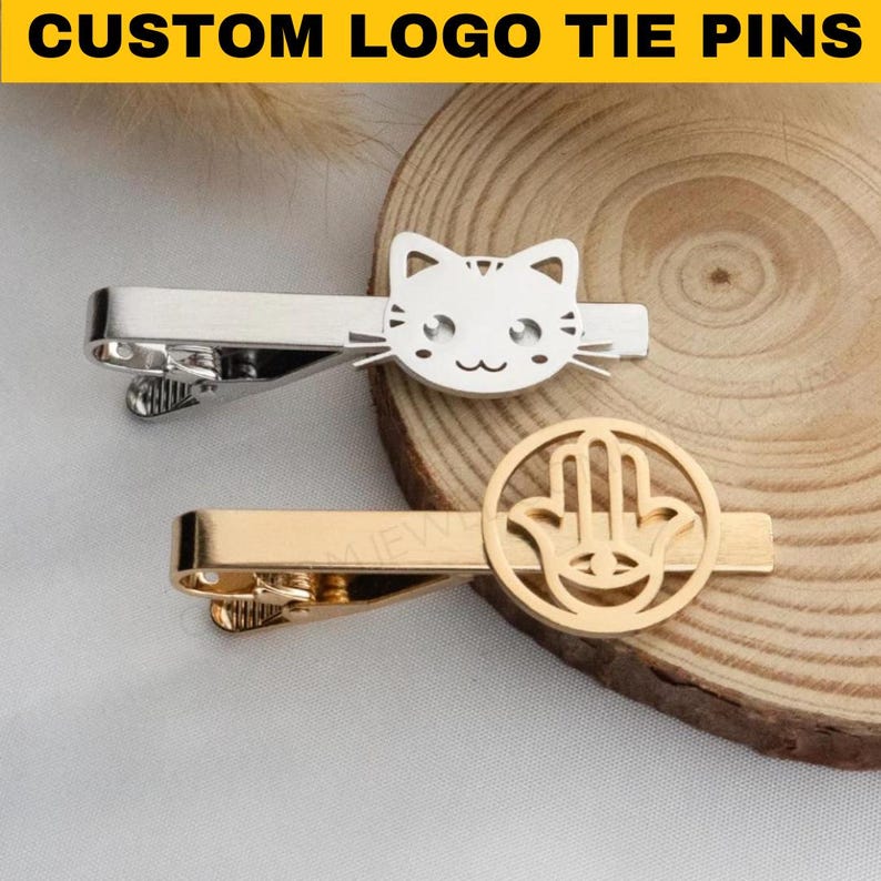 May include: Two custom tie pins. One is silver with a cat face design, the other is gold with a Hamsa hand symbol. The text "CUSTOM LOGO TIE PINS" is at the top of the image.