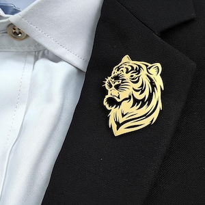 May include: A gold-colored metal tiger brooch pinned to a black blazer. The brooch features a detailed tiger head design, with black stripes and facial features. The blazer is worn over a white dress shirt.