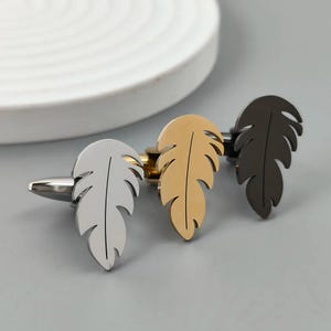 Puede incluir: Tres gemelos con forma de pluma en plata, oro y negro. Los gemelos son de metal y tienen un acabado pulido. El diseño de la pluma es detallado y elegante, adecuado para ocasiones formales.