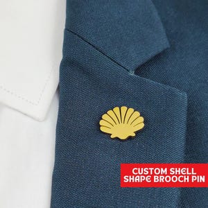 May include: A gold-coloured shell-shaped brooch pinned to a dark blue blazer. The brooch is a custom design, as indicated by the text "CUSTOM SHELL SHAPE BROOCH PIN" in a red banner at the bottom. A white shirt collar is visible in the background.