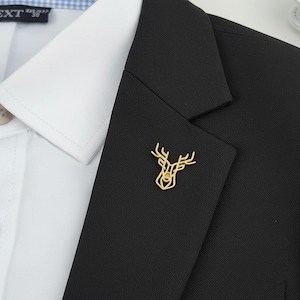 Könnte beinhalten: Goldfarbene Rehkopf-Anstecknadel auf einem schwarzen Blazer. Der Pin ist ein minimalistisches Design, das die Umrisse eines Rehkopfes mit Geweih zeigt. Ein weißes Hemd mit Kragen ist darunter sichtbar.